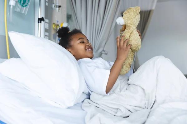 ハッピーアフリカ系アメリカ人少女患者は 病院の患者室でテディベアと遊んでベッドに横たわっている 小児期 医学および医療 変更されていない — ストック写真