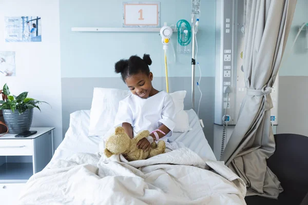 ハッピーアフリカ系アメリカ人少女患者は 病院の患者室でテディベアと遊んでベッドに横たわっている 小児期 医学および医療 変更されていない — ストック写真
