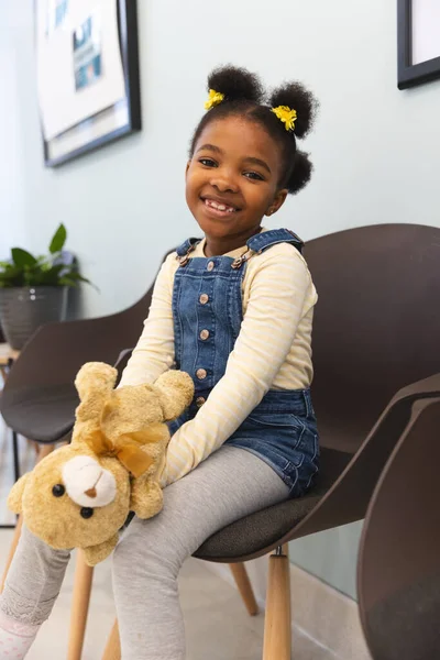 テディベアを抱えているアフリカ系アメリカ人の少女の肖像画は 病院で待合室に座っている 小児期 医学および医療 変更されていない — ストック写真