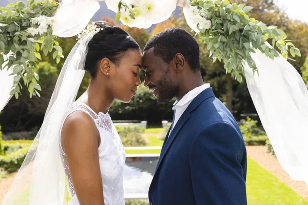 Happy african american bride and groom touching heads under wedding arch at ceremony in sunny garden. Marriage, romance, summer, tradition, ceremony and lifestyle, unaltered.