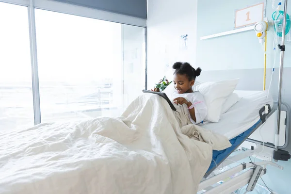 アフリカ系アメリカ人少女患者が病院の患者室でタブレットを使用してベッドに横たわっている 小児期 コミュニケーション 変わらない — ストック写真
