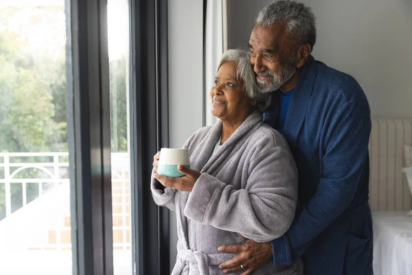 快乐的老夫妇穿着浴衣 带着杯子咖啡从窗户往外看 老年生活方式 退休和家庭生活 — 图库照片