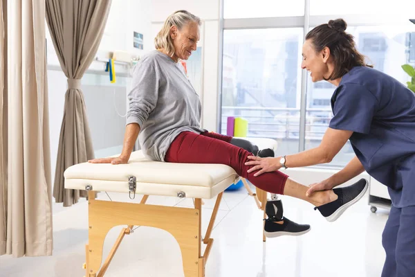 Caucasian female physiotherapist and senior woman with artificial leg stretching at hospital. Hospital, disability, physiotherapy, work, medicine and healthcare, unaltered.