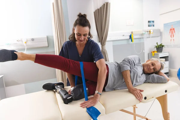Caucasian female physiotherapist and senior woman with artificial leg using exercise band stretching. Hospital, disability, physiotherapy, work, medicine and healthcare, unaltered.
