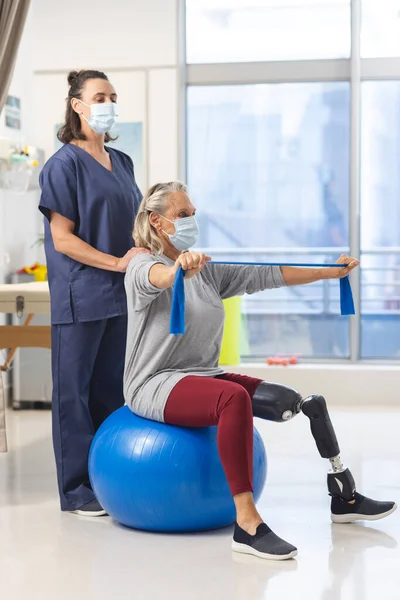 Caucasian physiotherapist and senior woman with artificial leg with face masks using exercise band. Hospital, hygiene, disability, physiotherapy, work, medicine and healthcare, unaltered.