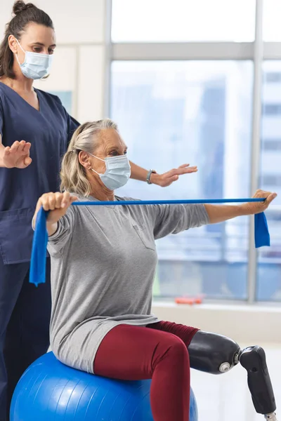 Caucasian physiotherapist and senior woman with artificial leg with face masks using exercise band. Hospital, hygiene, disability, physiotherapy, work, medicine and healthcare, unaltered.