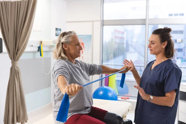 Caucasian female physiotherapist and senior woman with artificial leg using exercise band stretching. Hospital, disability, physiotherapy, work, medicine and healthcare, unaltered.