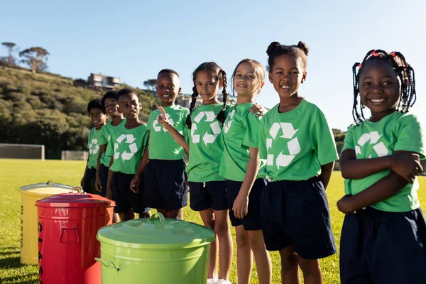 Portrait of happy diverse schoolchildren wearing green recycling tshirts at recycling bins. Recycling, environment, education, childhood, development, learning and school, unaltered.