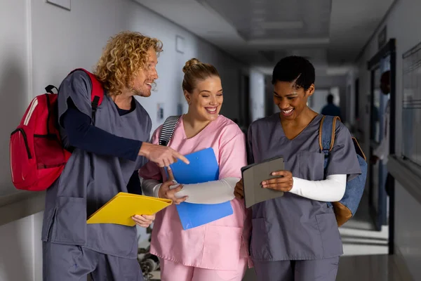 Happy diverse doctors wearing scrubs discussing work and using tablet in corridor at hospital. Hospital, communication, teamwork, medicine, healthcare and work, unaltered.