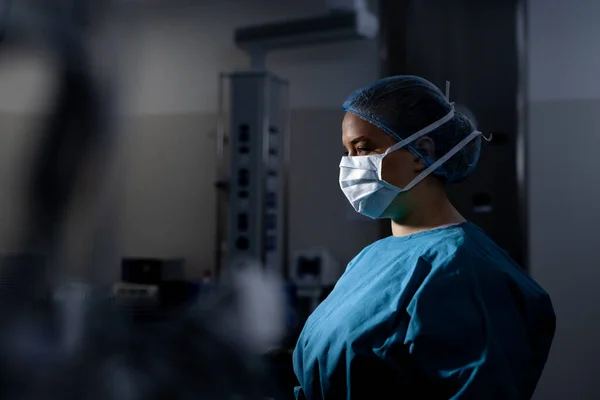 African american female surgeon wearing surgical gown and face mask in operating theatre at hospital. Hospital, surgery, hygiene, medicine, healthcare and work, unaltered.