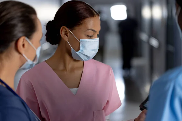 Diverse female doctors wearing face masks using tablet and talking in corridor at hospital. Hospital, communication, teamwork, medicine, healthcare and work, unaltered.
