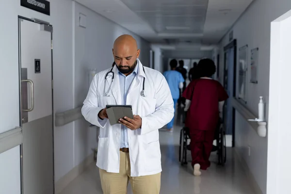 Biracial male doctor wearing lab coat using tablet in corridor at hospital. Hospital, communication, medicine, healthcare and work, unaltered.