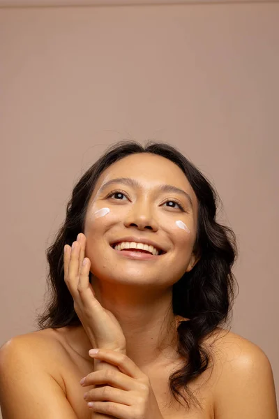Smiling asian woman with dark hair with skin cream on her cheeks touching face, copy space. Femininity, face, facial expressions, body, skin and beauty treatments, unaltered.