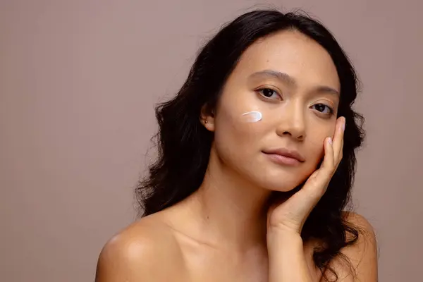 Asian woman with dark hair with skin cream on her cheek touching face with hand, copy space. Femininity, face, facial expressions, body, skin and beauty, unaltered.