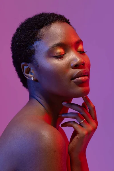 African american woman touching chin with eyes closed in red light on purple background. Femininity, face, facial expressions, body, skin, makeup, fashion and beauty, unaltered.