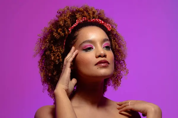 Biracial woman with pink eye shadow and lipstick touching face, purple background. Femininity, face, facial expressions, body, skin, makeup, fashion and beauty, unaltered.