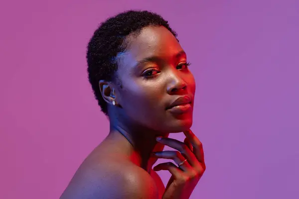 African american woman touching chin in red light on purple background. Femininity, face, facial expressions, body, skin, makeup, fashion and beauty, unaltered.