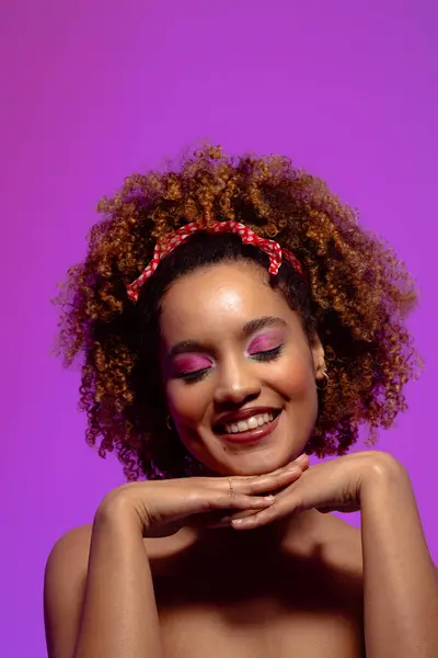 Smiling biracial woman with pink eye shadow, eyes closed and hands under chin, purple copy space. Femininity, face, facial expressions, body, skin, makeup, fashion and beauty, unaltered.