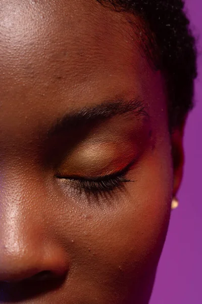 Close up of closed eye of african american woman with short hair on purple background. Femininity, face, facial expressions, body, skin, makeup, fashion and beauty, unaltered.