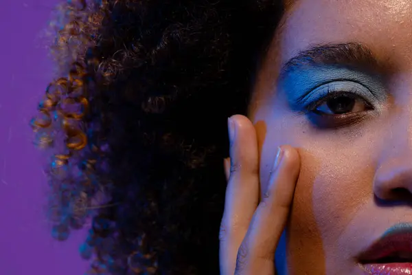 Half portrait of biracial woman with curly hair and blue eye shadow touching face with hand. Femininity, face, facial expressions, body, skin, makeup, fashion and beauty, unaltered.
