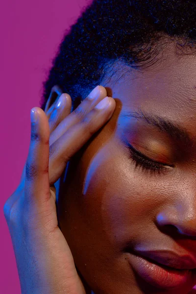 African american woman with eye closed touching head in blue light on purple background. Femininity, face, facial expressions, body, skin, makeup, fashion and beauty, unaltered.