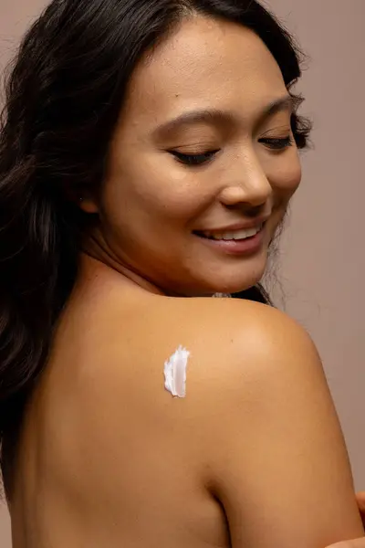 Smiling asian woman with dark hair with skin cream on her bare shoulder. Femininity, face, facial expressions, body, skin and beauty treatments, unaltered.