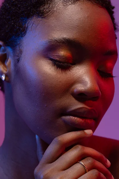 Clos up of african american woman with eyes closed holding chin in red and blue light. Femininity, face, facial expressions, body, skin, makeup, fashion and beauty, unaltered.