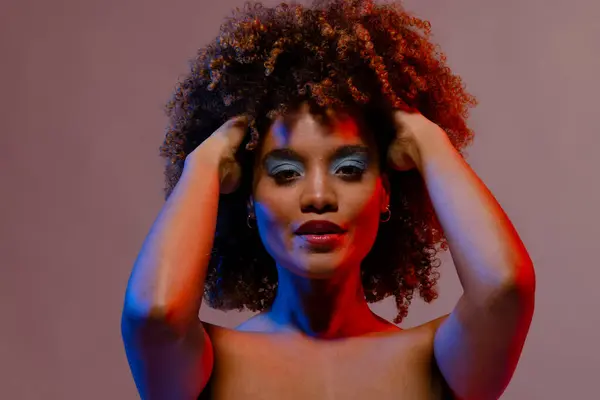 stock image Biracial woman in blue eye shadow with hands in curly hair in blue and red light. Femininity, face, facial expressions, body, skin, makeup, fashion and beauty, unaltered.