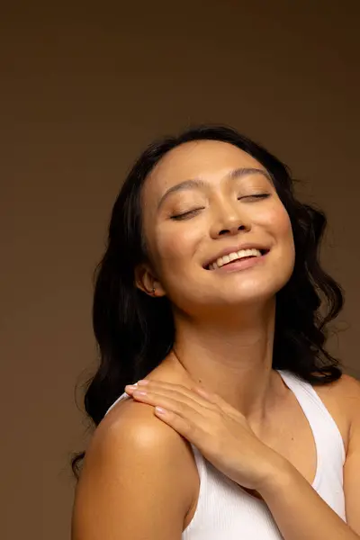 Happy asian woman with dark hair, with hand on shoulder and eyes closed, copy space. Femininity, face, facial expressions, body, skin and beauty, unaltered.