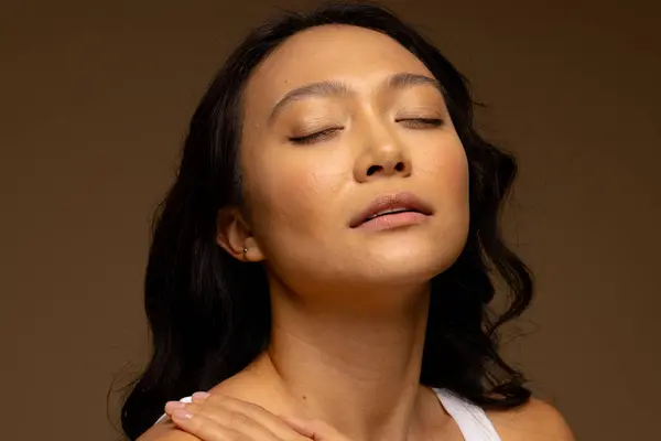 Asian woman with dark hair, with hand on shoulder and eyes closed. Femininity, face, facial expressions, body, skin and beauty, unaltered.