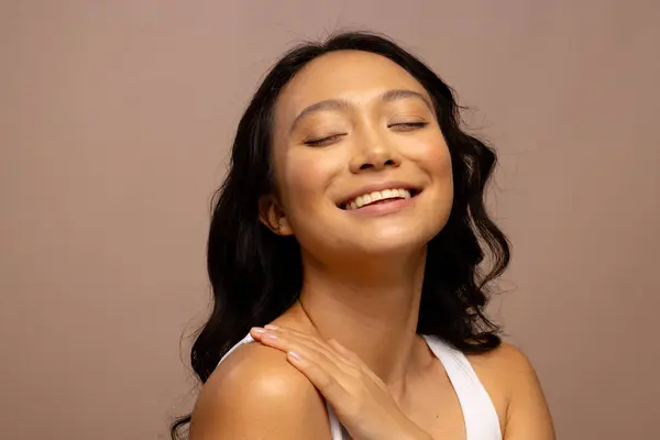 Happy asian woman with dark hair, with hand on shoulder and eyes closed. Femininity, face, facial expressions, body, skin and beauty, unaltered.