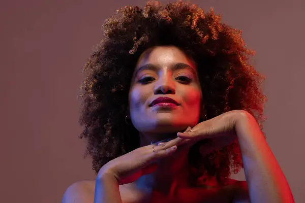 Smiling biracial woman with hands under chin in blue and red light. Femininity, face, facial expressions, body, skin, makeup, fashion and beauty, unaltered.