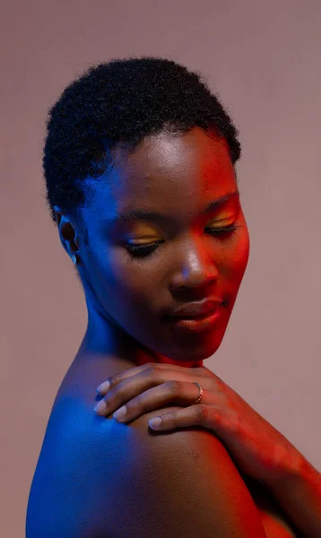 African american woman with short hair looking down with hand on shoulder in blue and red light. Femininity, face, facial expressions, body, skin, makeup, fashion and beauty, unaltered.