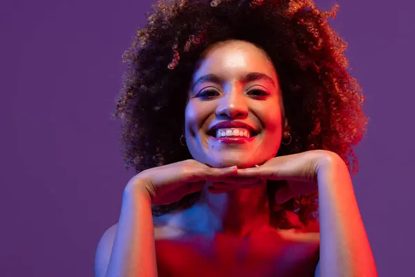 Happy biracial woman with hands under chin in blue and red light on purple background. Femininity, face, facial expressions, body, skin, makeup, fashion and beauty, unaltered.
