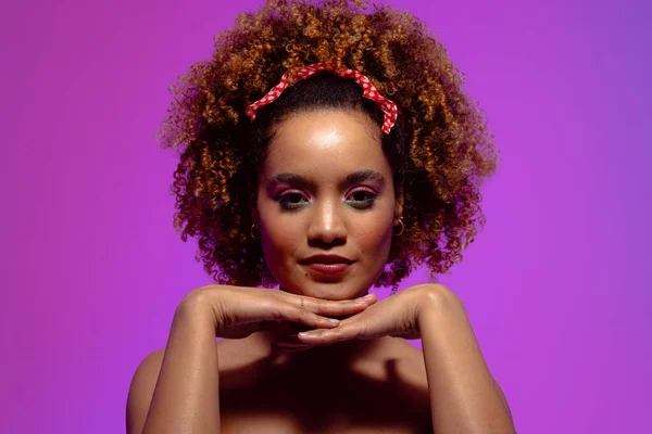 Biracial woman with curly hair, pink eye shadow and lipstick, hands under chin on purple background. Femininity, face, facial expressions, body, skin, makeup, fashion and beauty, unaltered.