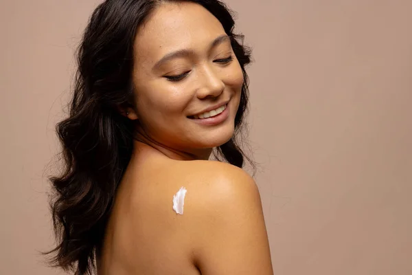 Smiling asian woman with dark hair with skin cream on her bare shoulder, copy space. Femininity, face, facial expressions, body, skin and beauty treatments, unaltered.