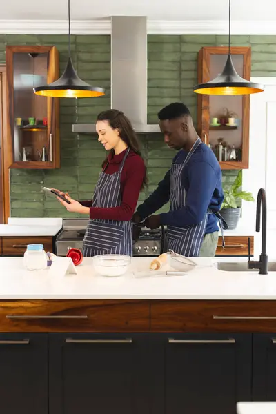 Happy diverse couple baking together in kitchen, using tablet at home. Lifestyle, togetherness, relationship, baking, recipe, communication and domestic life, unaltered.