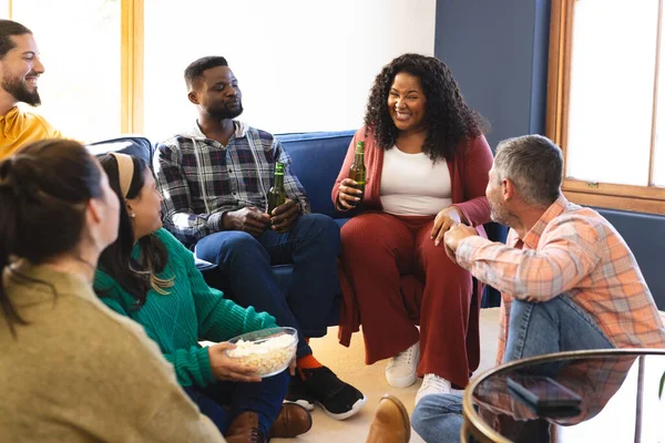 Stock image Happy diverse male and female friends talking, eating popcorn and drinking beer at home. Lifestyle, friendship, relaxation, free time and domestic life, unaltered.