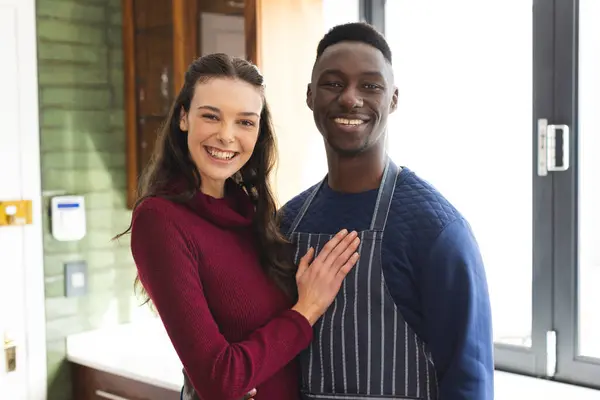 Portrait of happy diverse couple embracing and smiling in kitchen at home. Lifestyle, togetherness, relationship, cooking and domestic life, unaltered.
