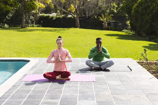 Diverse couple practicing yoga and meditating next to swimming pool in garden. Lifestyle, togetherness, relationship, relaxation and domestic life, unaltered.
