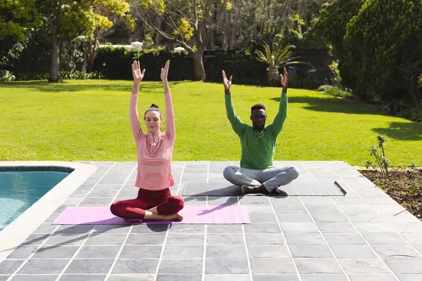 Diverse couple practicing yoga and meditating next to swimming pool in garden. Lifestyle, togetherness, relationship, relaxation and domestic life, unaltered.