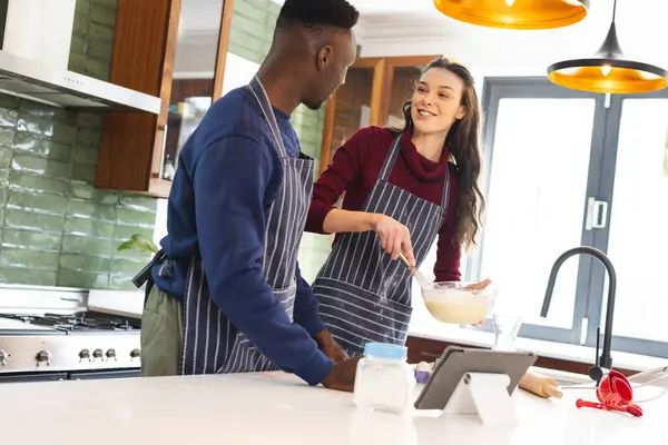 Happy diverse couple baking together in kitchen, using tablet at home. Lifestyle, togetherness, relationship, baking, recipe, communication and domestic life, unaltered.