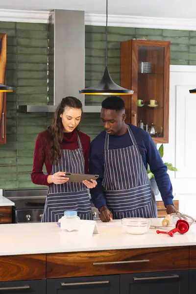 Happy diverse couple baking together in kitchen, using tablet at home. Lifestyle, togetherness, relationship, baking, recipe, communication and domestic life, unaltered.