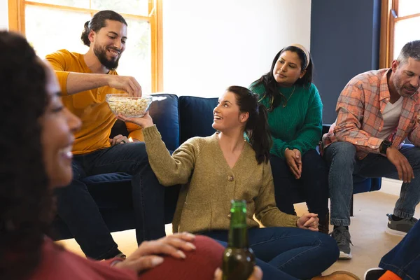 Happy diverse male and female friends talking, eating popcorn and drinking beer at home. Lifestyle, friendship, relaxation, free time and domestic life, unaltered.