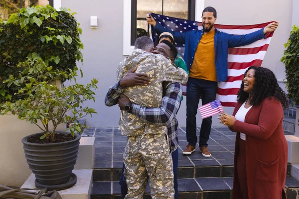Happy diverse friends with flags welcoming home male soldier friend. Military service, returning home, celebration, patriotism and friendship, unaltered.