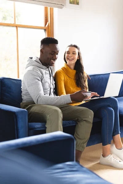 Stock image Happy diverse couple sitting on sofa using laptop for online shopping at home. Lifestyle, togetherness, relationship, online shopping, communication and domestic life, unaltered.