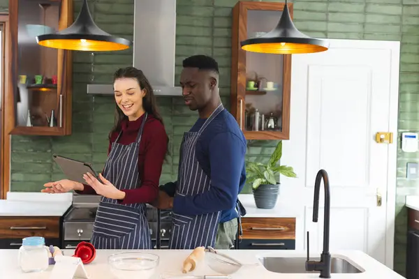 Happy diverse couple baking together in kitchen, using tablet at home, copy space. Lifestyle, togetherness, relationship, baking, recipe, communication and domestic life, unaltered.