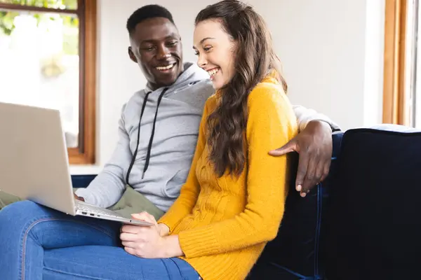 stock image Happy diverse couple sitting on sofa using laptop at home. Lifestyle, togetherness, relationship, communication and domestic life, unaltered.