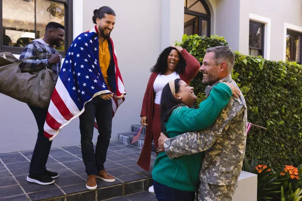Happy diverse friends with flags welcoming home male soldier friend. Military service, returning home, celebration, patriotism and friendship, unaltered.