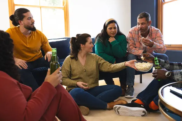 Happy diverse male and female friends talking, eating popcorn and drinking beer at home. Lifestyle, friendship, relaxation, free time and domestic life, unaltered.